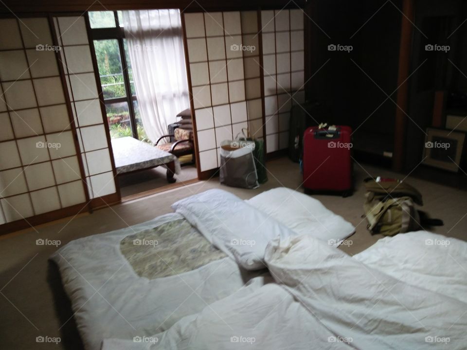 a Japanese tradisional bed room. white bed matras, pillow and blanket on the wooden floor and suitcases. sliding door and a table in seperate room facing to the garden.