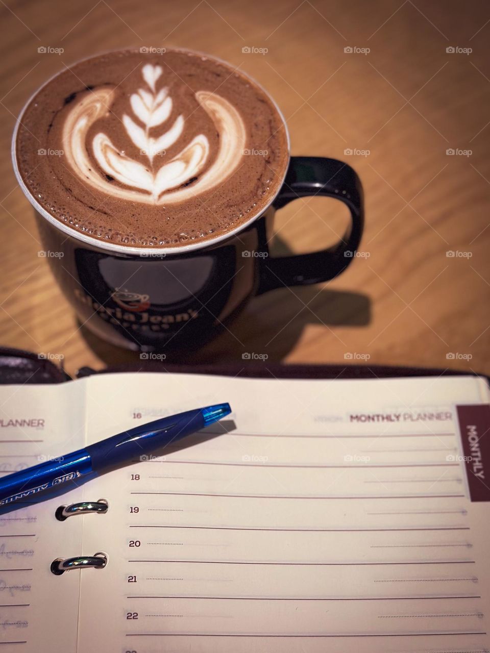 A coffee cup with latte art next to an organiser with a pen