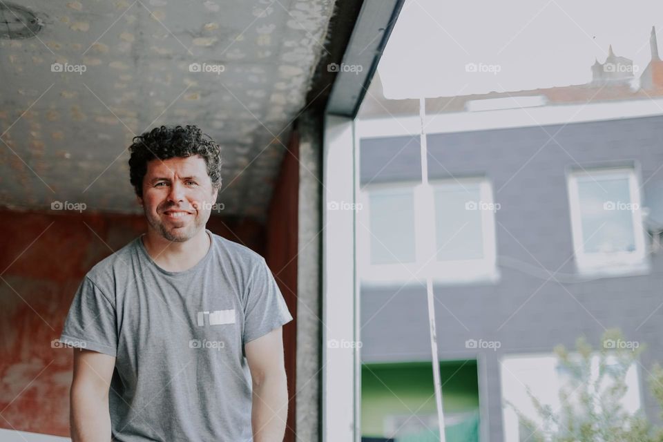 Portrait of a young handsome Caucasian man brunet with curly hair in a gray T-shirt with a happy smile on his face looks into the camera and stands near the window in the room where there is a construction repair on the installation of window frames