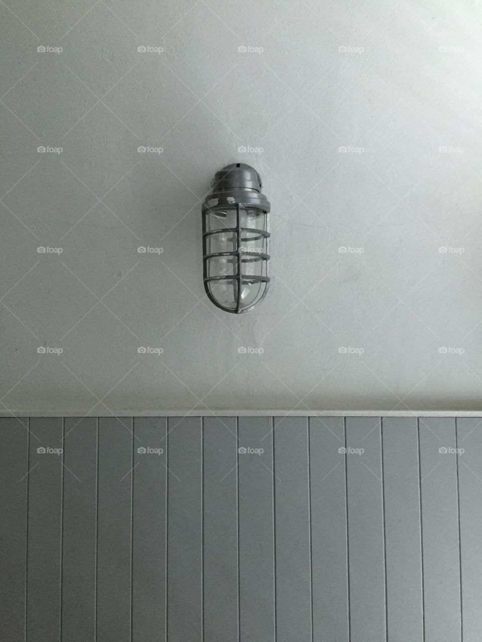 A light bulb protected by a metallic frame on a plain grey wall. The lower part of the wall has a gray wooden pattern.