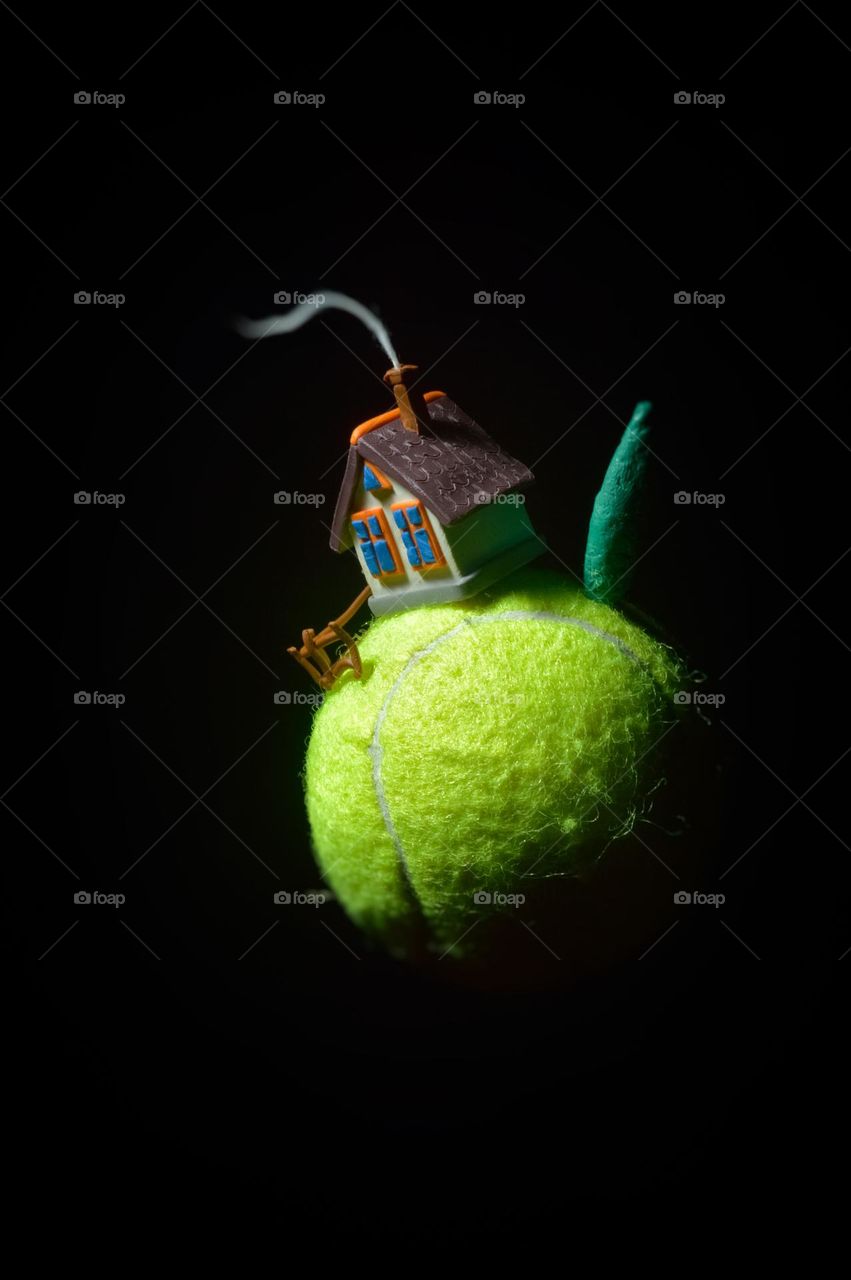 volumetric plasticine miniature. small planet tennis ball. there is a plasticine house on the ball. a one-story house with smoke from a chimney in complete darkness. a small fence and a tree.