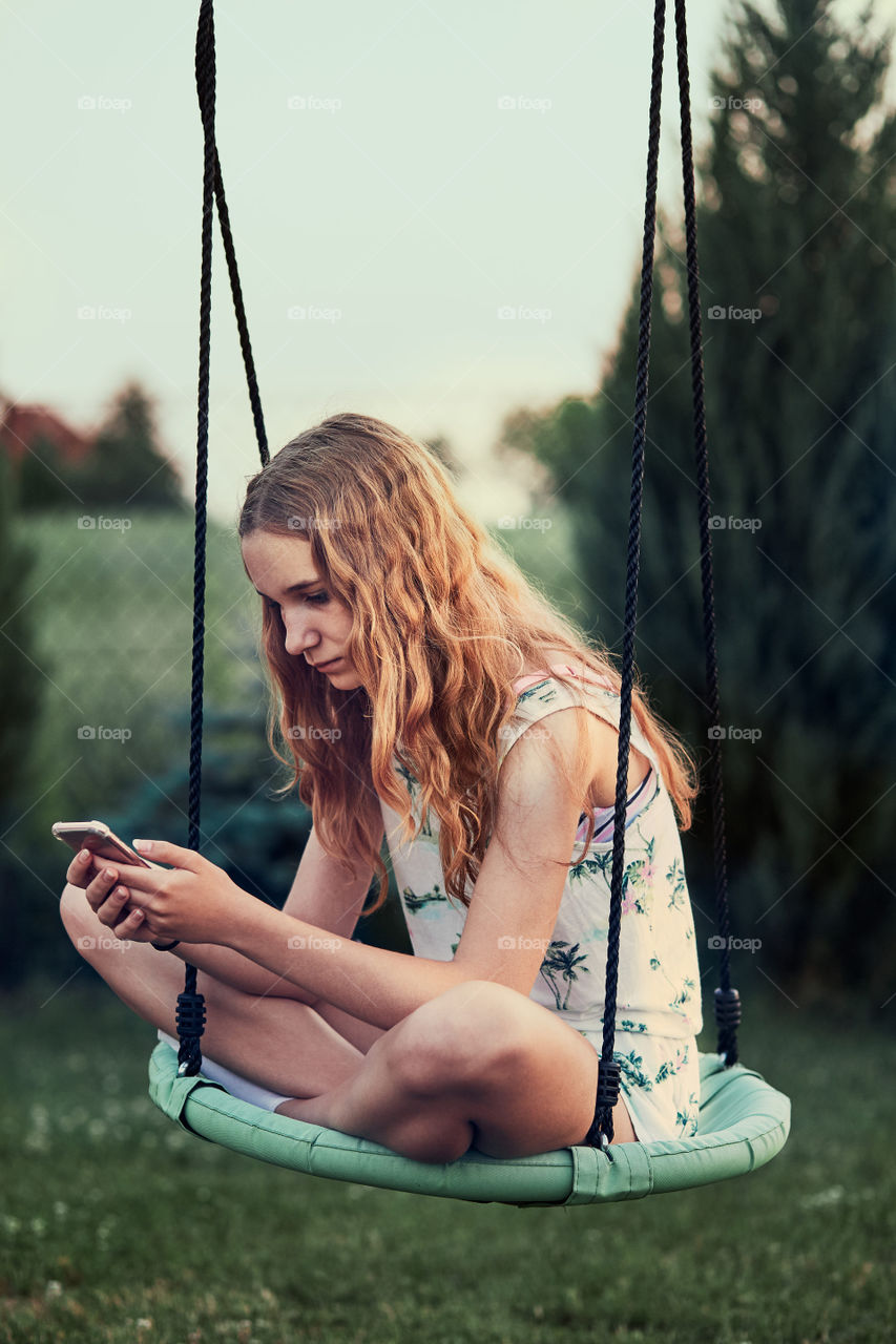 Young woman using mobile phone smartphone sitting on swing in a backyard. Candid people, real moments, authentic situations