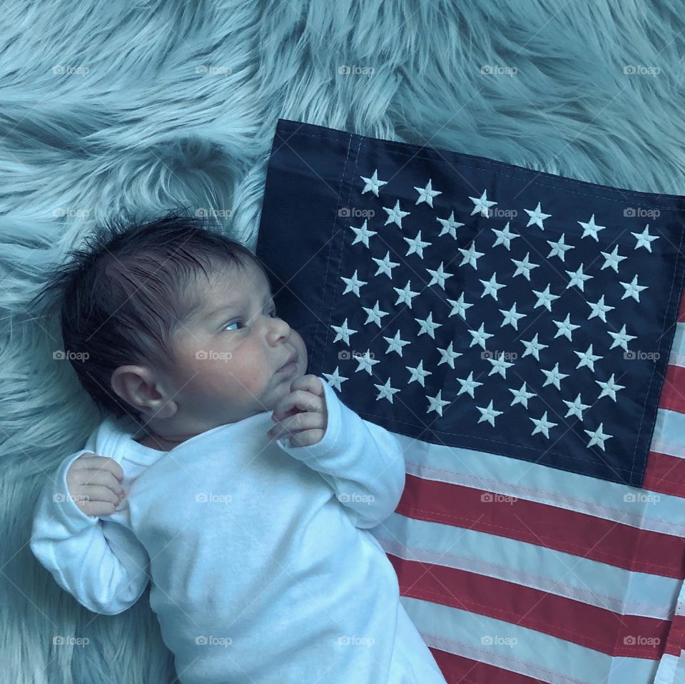 Baby looks curiously at the United States flag, baby with American flag, patriotic baby with flag, baby looking at American flag, United States of America flag with infant, newborn baby with flag 