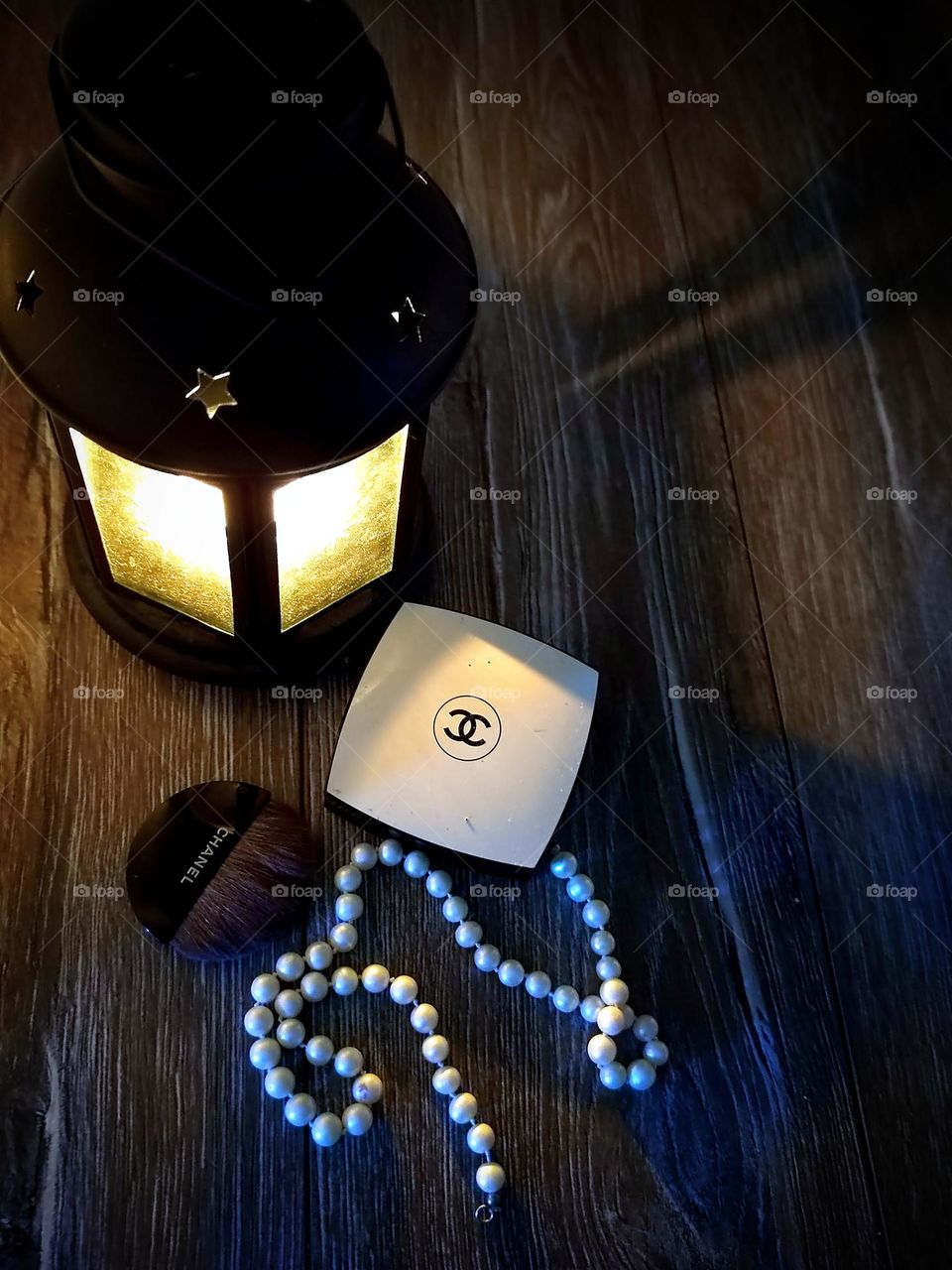 A lantern is lit on a wooden background, the light from which falls on a box with the Chanel logo, on a white string of pearls and a tassel with the inscription "CHANEL". Powder Chanel Les Beiges