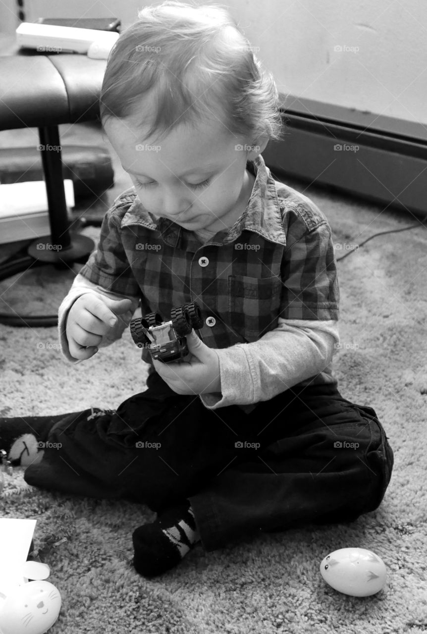 A toddler boy sitting on the carpeted floor plays with a toy truck figuring out how all the moving parts work. 