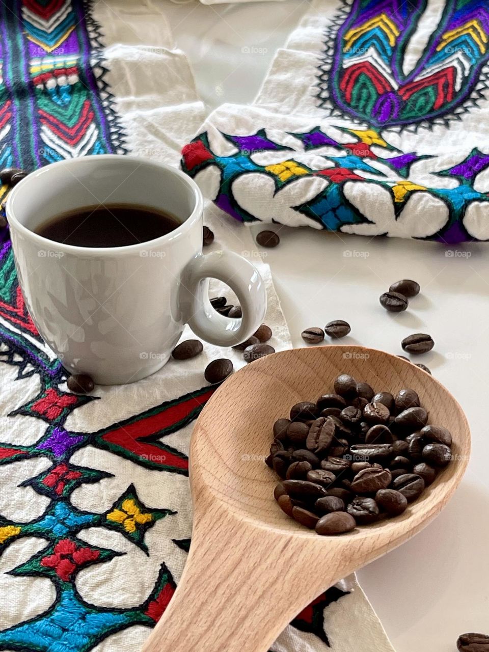 coffee theme composition: white coffee cup containing liquid coffee and coffee beans on white surface and inside a natural wooden spoon, on hand-embroidered Eritrean drape with typical ethnic embroideries