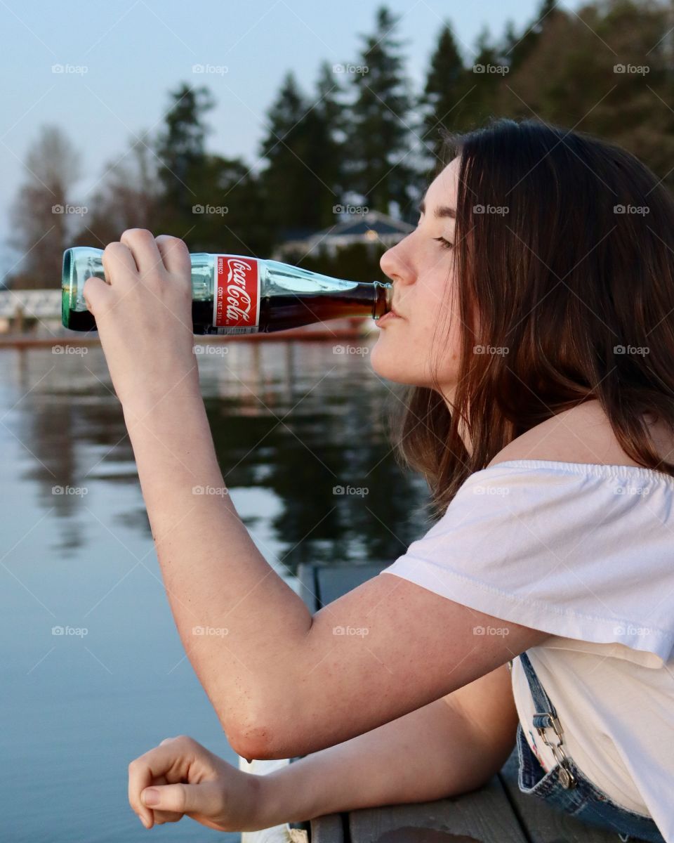 A young woman enjoys a refreshing bottle of Coca Cola while relaxing on a dock overlooking calm blue waters