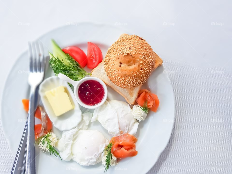 Top view to white plate with breakfast made by eggs, salted salmon fish, butter, vegetables, cream cheese, bread.