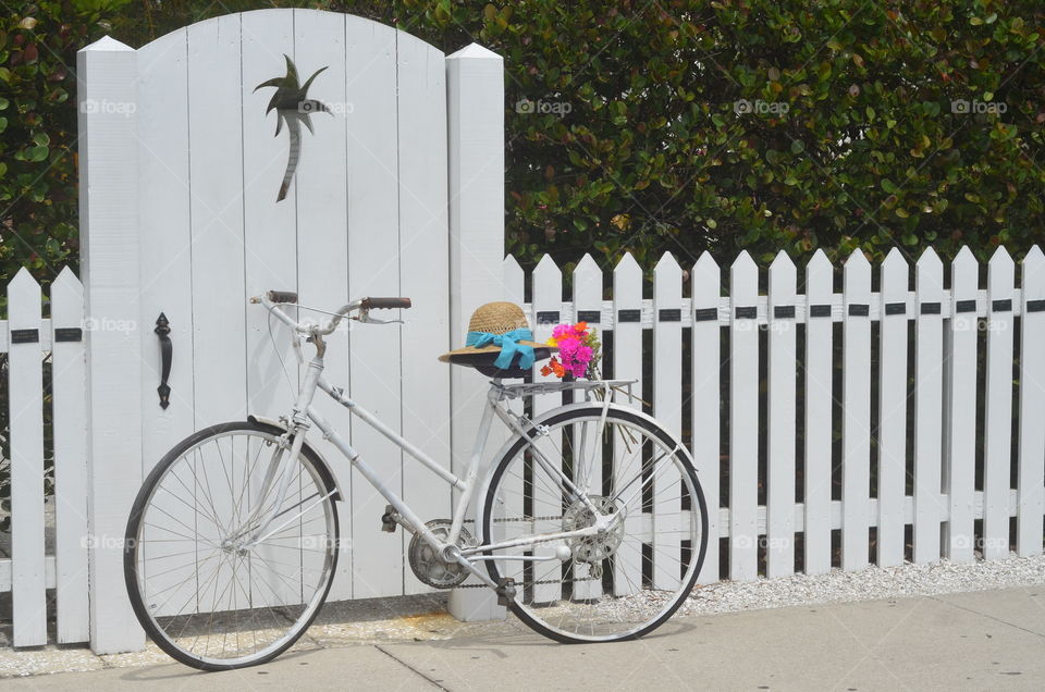 Classic Vintage White Bike Against White Picket Fence With Vibrant Flowers and A Ladies Sunhat