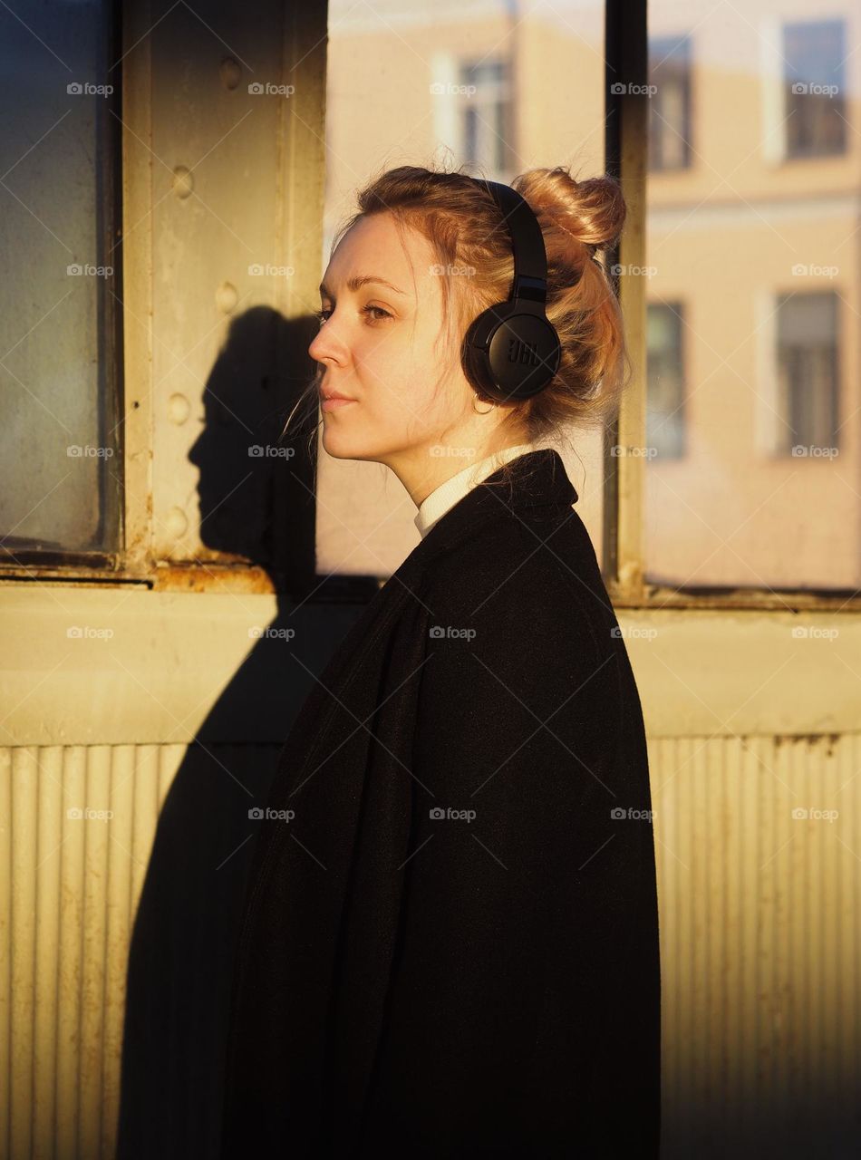 Young blonde woman in headphones listen music and looking at window in sunny day, portrait of woman.