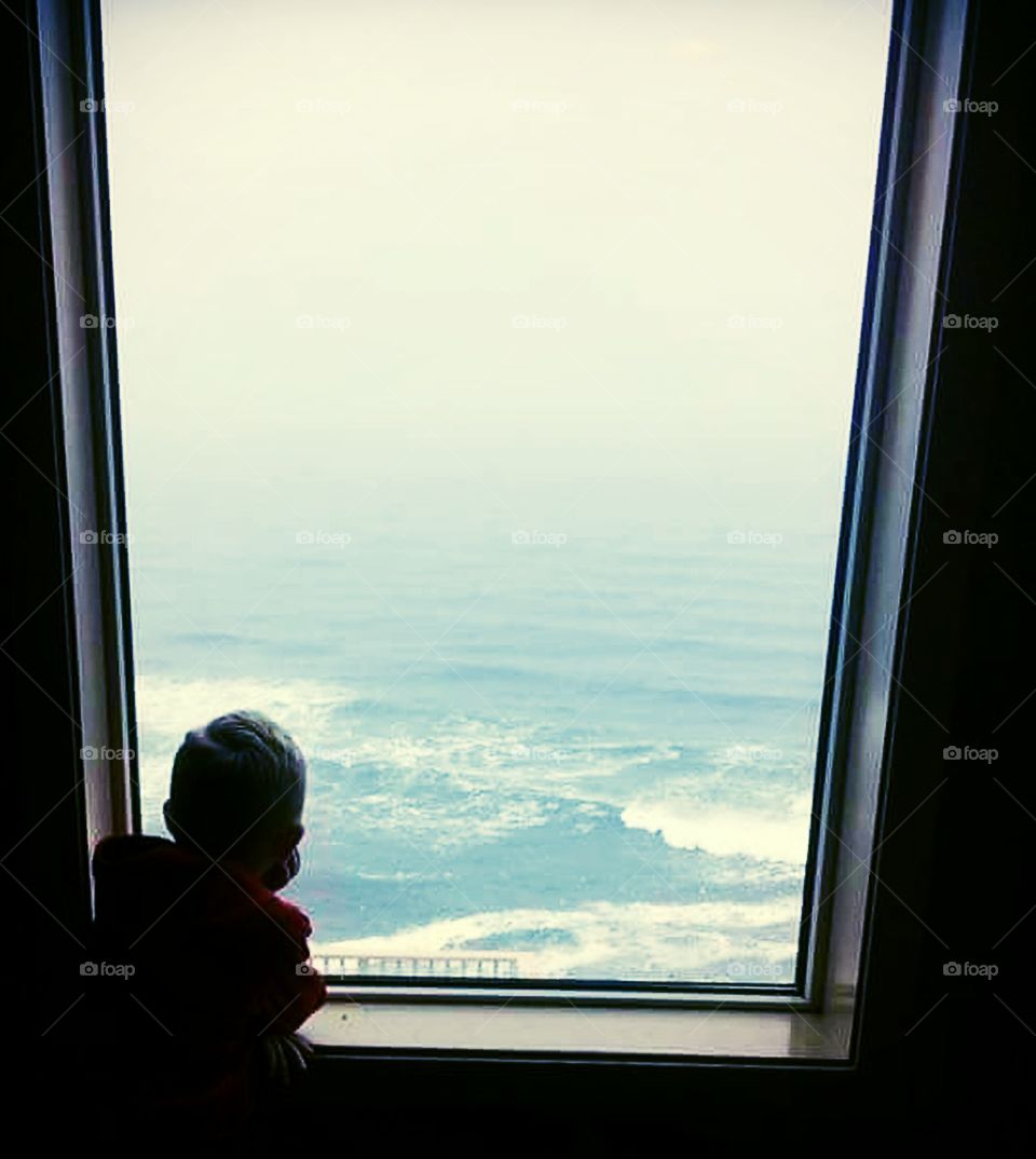 Boy watches through a window with wonder, as waves roll in on Oregon coast cliffsides.