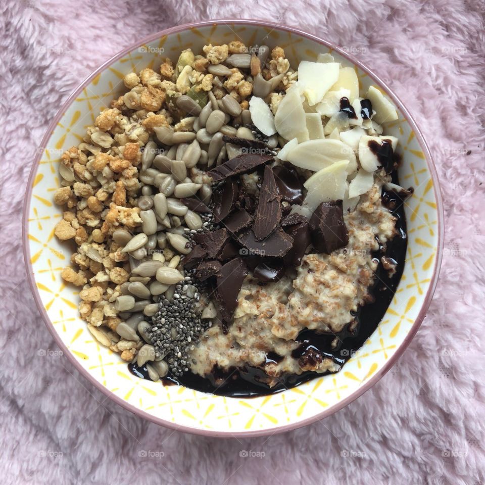 Oats in a bowl topped with granola chocolate Nuts and seeds in a pink textured background 