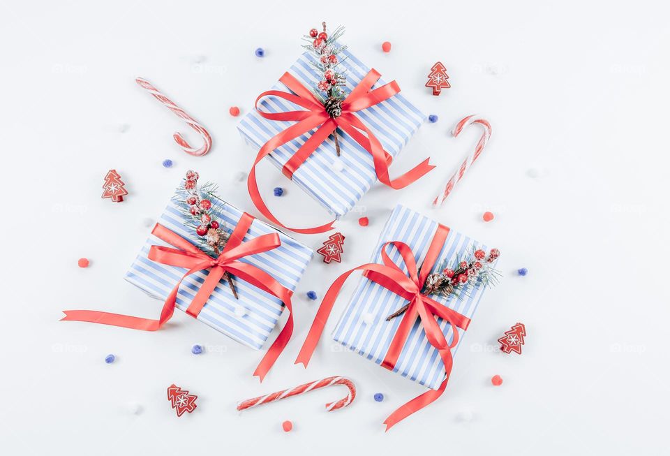 Three blue striped gift boxes tied with a red ribbon, lollipops, decorative fir branches, fluffy snow and Christmas confetti lie in the center on a white background, flat lay close-up. Holiday gifts concept.