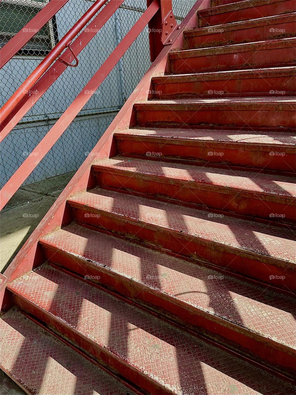The late afternoon sun casts strong shadows across the stepped structures of the red metal staircase of the “Pulaski Bridge” at “Newtown Creek” in LIC, Queens and adds life and movement to its minimalist design. 2024. Hypnotic Productions