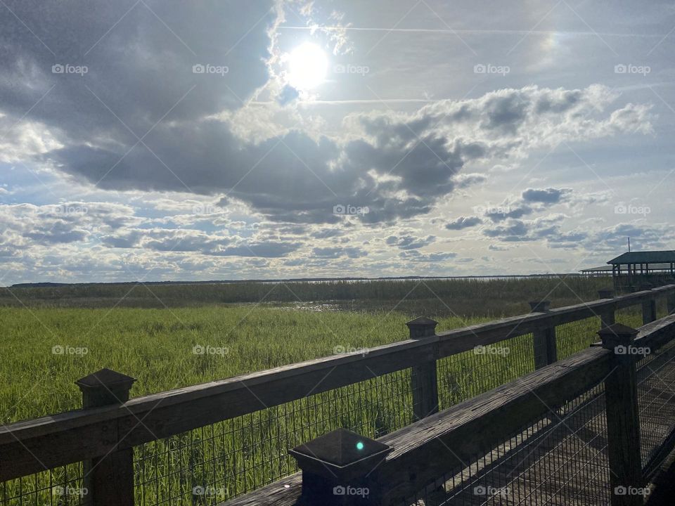 A wooden bridge  across a grassy marsh under a sky with the sun peeping out of rain clouds