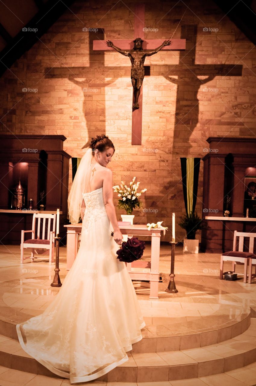 Breathtaking image of a beautiful bride captured at the alter moments before her wedding in a Catholic Church with unique lighting creating shadows behind a hanging crucifix giving an illusion of the trinity being present at mass 