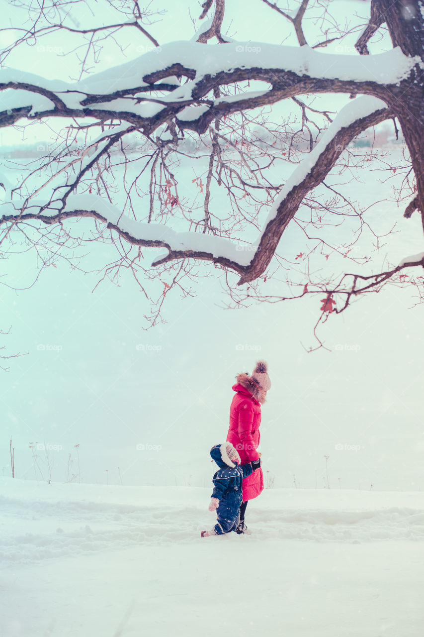 Mother and her little daughter are spending time together walking outdoors in forest in winter while snow falling going through deep snow enjoying wintertime