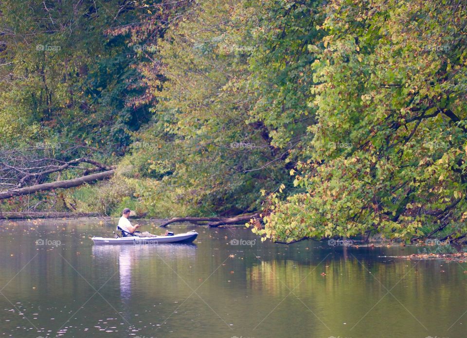 Man in a small boat fishing, trees reflecting in the water 