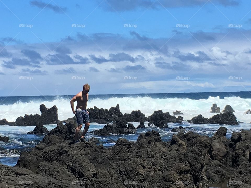 Young man walking on rocks with blue skies and waves breaking behind him in Maui, Hawaii.