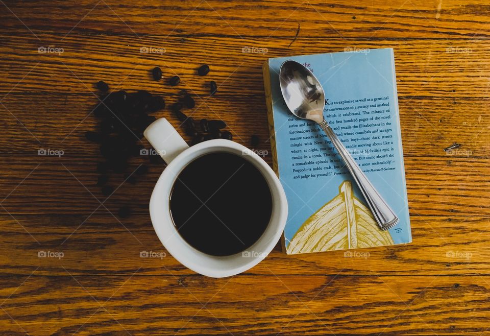 Black coffee in a white mug sitting on a wood table next to an old, vintage book with a silver spoon and dark roast coffee beans