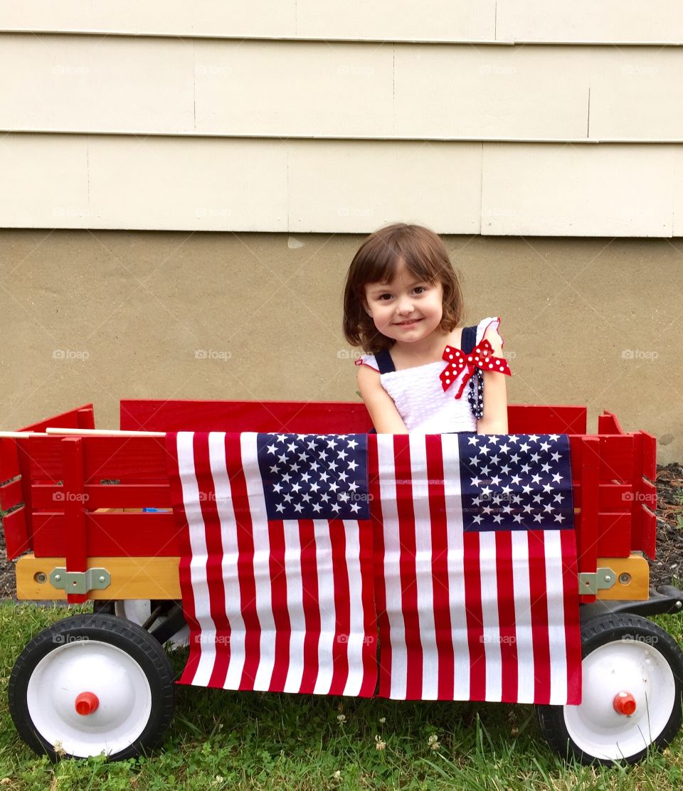 A little girl sitting on wooden cart with American flag