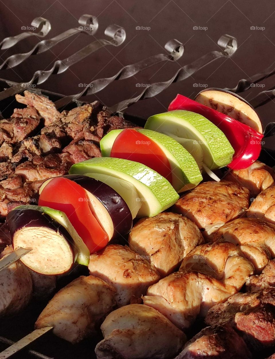 Skewers on the grill with coals.  Pieces of fried meat on skewers.  One vegetable skewer: tomato rings, zucchini rings and eggplant rings