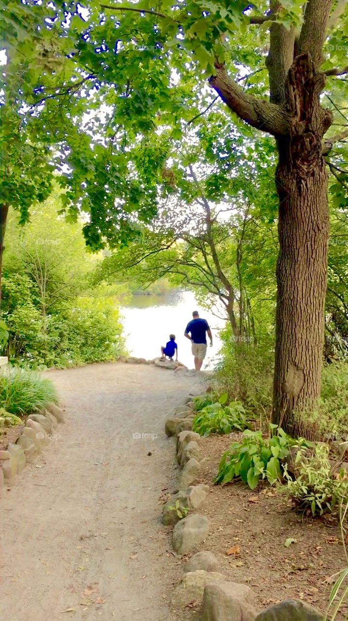 A man with a young boy exploring nature via dirt path through the woods at edge of river, at Roger Williams Park Zoo in Rhode Island, during summer