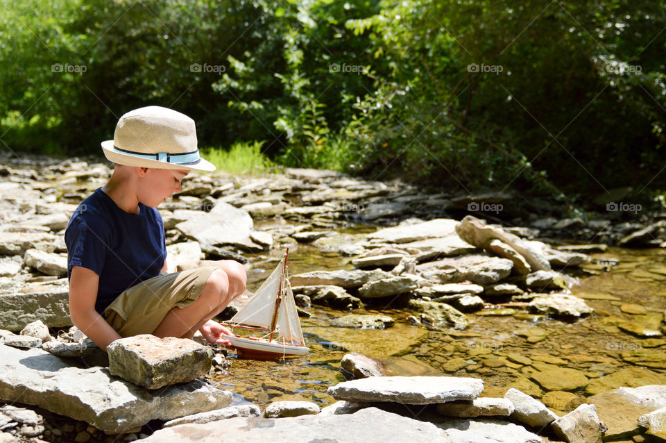 Boy playing with a toy sailboat in a creek during the summer