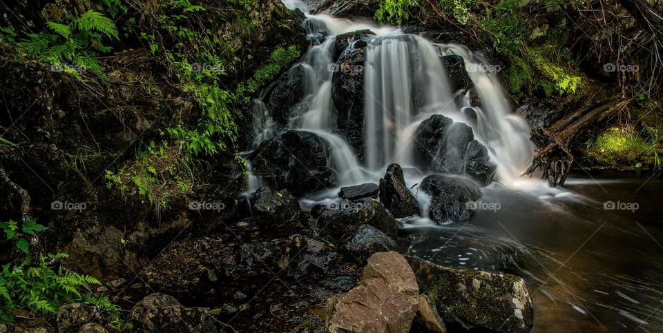 Small waterfalls with black stones and green leaves, in forest, summer, long exposure, horizontal