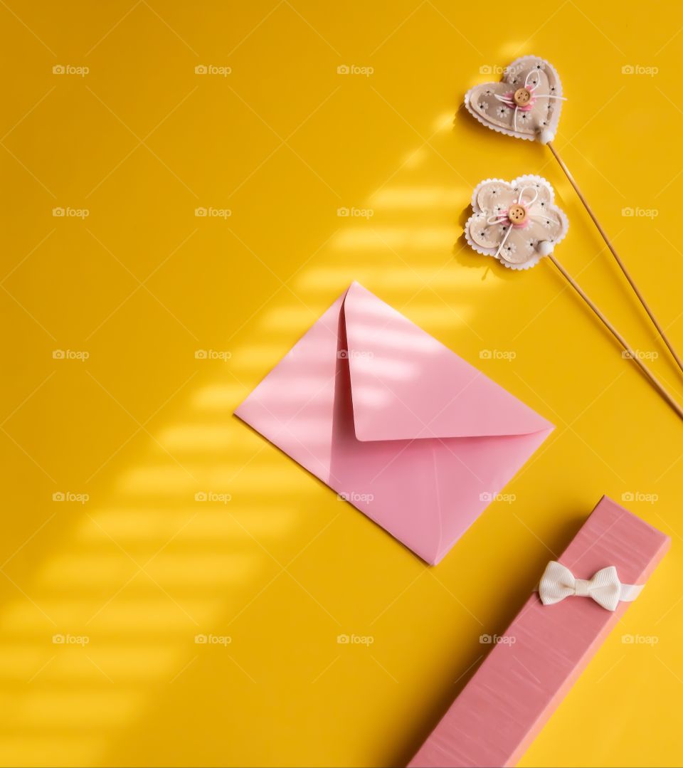 Still life made by pink gift box, handmade fabric decoration and envelope on yellow background. Top view.