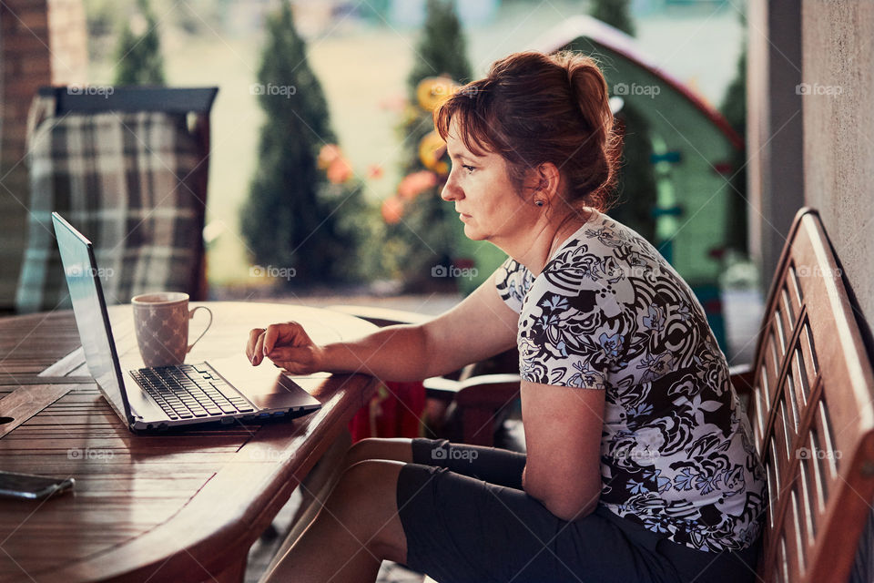 Woman working at home, using portable computer, sitting on patio on summer day. Candid people, real moments, authentic situations