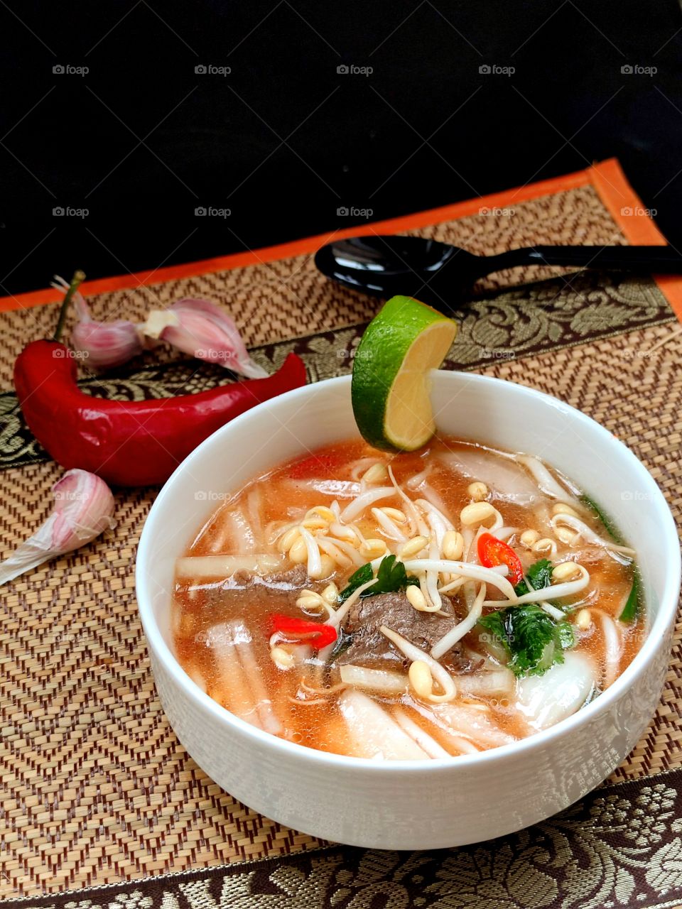 Thai beef soup.  In a white plate, pieces of beef, millet sprouts, noodles, pieces of red chili and pieces of garlic.  A slice of lime weighs on a plate.  Garlic cloves and chili peppers next to the plate
