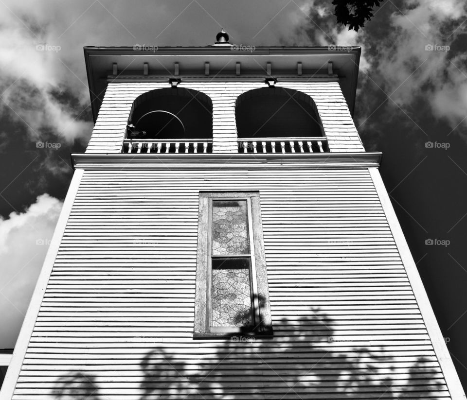 Old wood slat hundred year old country church place of worship building with bell tower and stained glass window with shadow of tree and cloudy sky in black and white perspective looking up