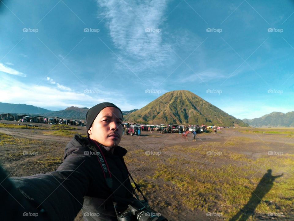 Selfie photo of an Asian man wearing warm clothes with a backdrop of views of the Bromo mountain area and blue sky during the day.
