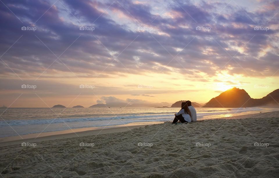 “The two sit alone on the beach and watch the sunset.
They look out over the water, the sun  setting behind the hills to their right."