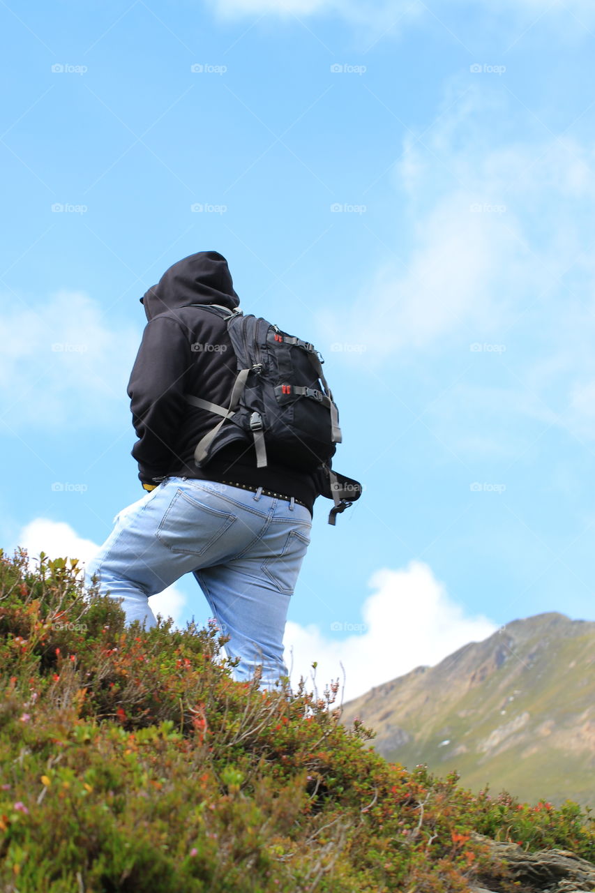 A man in blue jeans, a sweatshirt with a hood and a backpack walks along the Alpine mountains