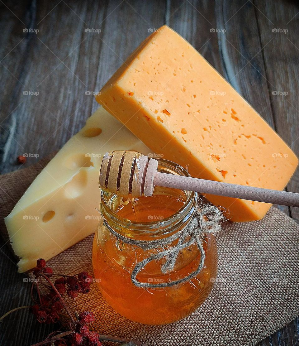 Food.  On a wooden background lies burlap, on which lie two types of cheese: white "Masdam" and yellow "Cheddar".  In the foreground is a jar of honey on which lies a spiral wooden spoon.  Honey is dripping from the spoon.