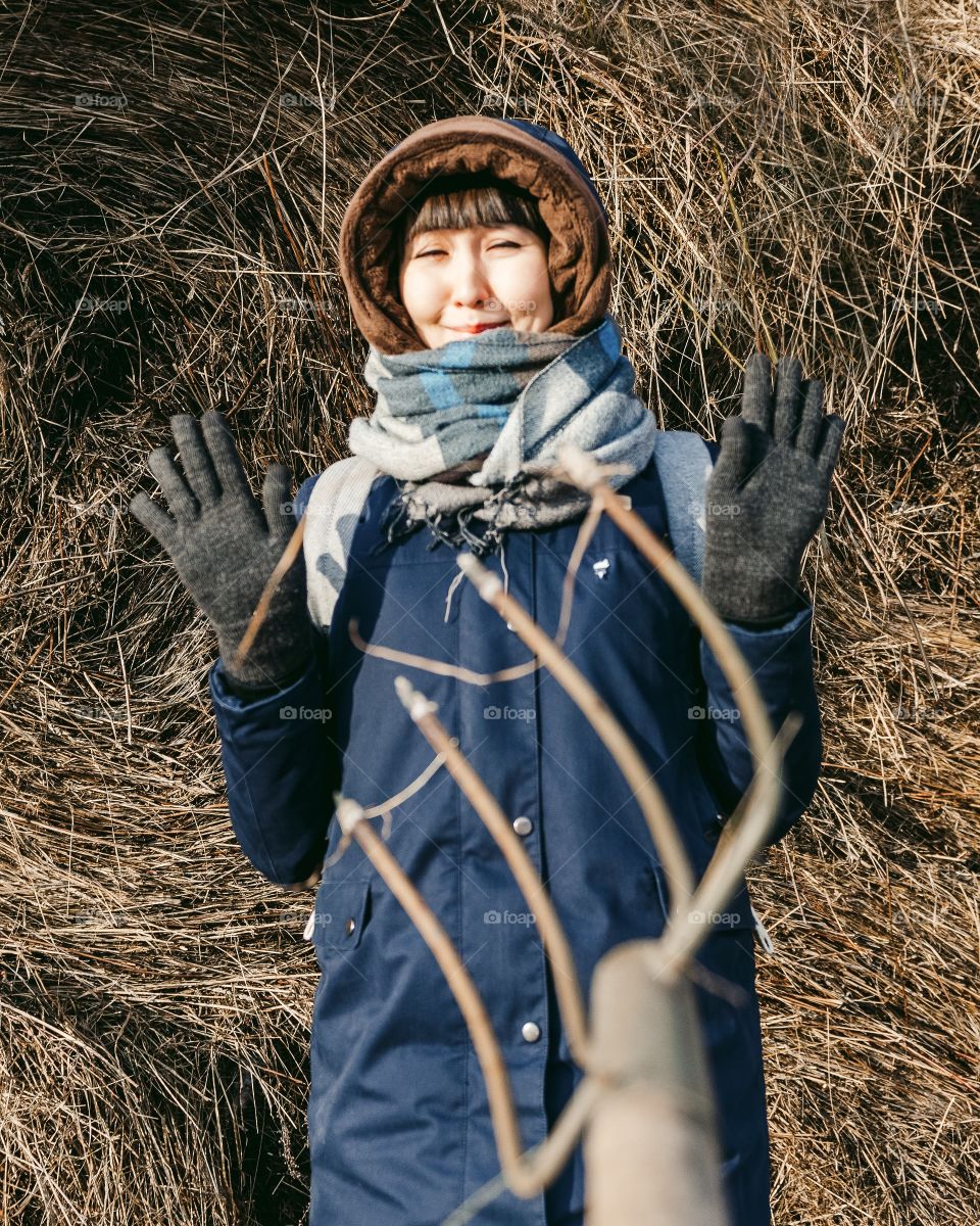 Cute asian girl standing in scarf and winter jacket near stack of hay threaten pitchfork