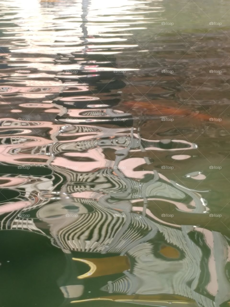 Shadows from objects above reflected onto the undulating surface of the water distorting them into facinating, patterns, shapes and colors. A solitary orange Koi fish swimming centre right just below water surface.