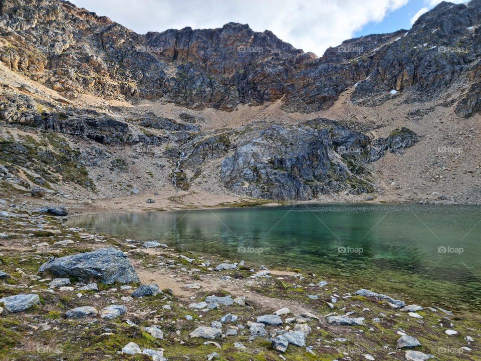shore of a green lagoon, sourrounded by mountains. Cloudy sky.