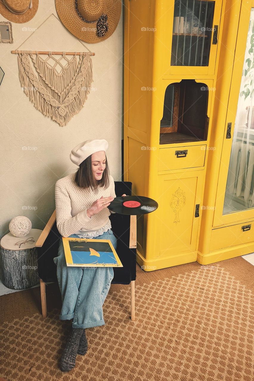 Portrait of beautiful young girl,  lady in beret sitting at cozy home in armchair near yellow vintage cabinet.  Vinyl music.