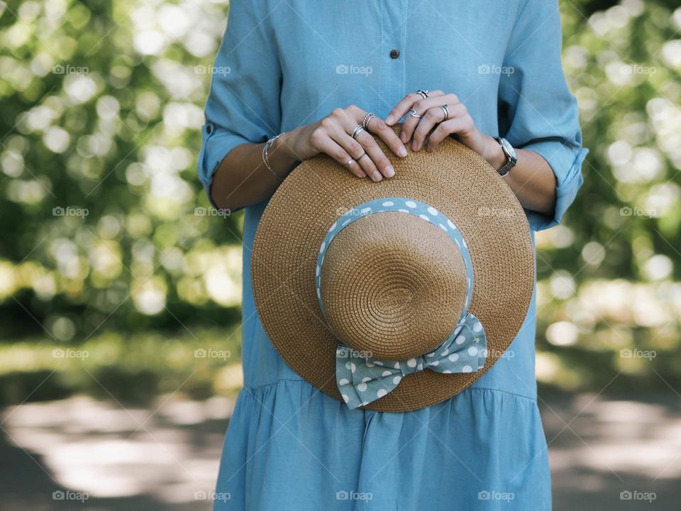 Unidentified young girl in a blue dress holding a straw hat standing in a green park on a sunny summer afternoon 