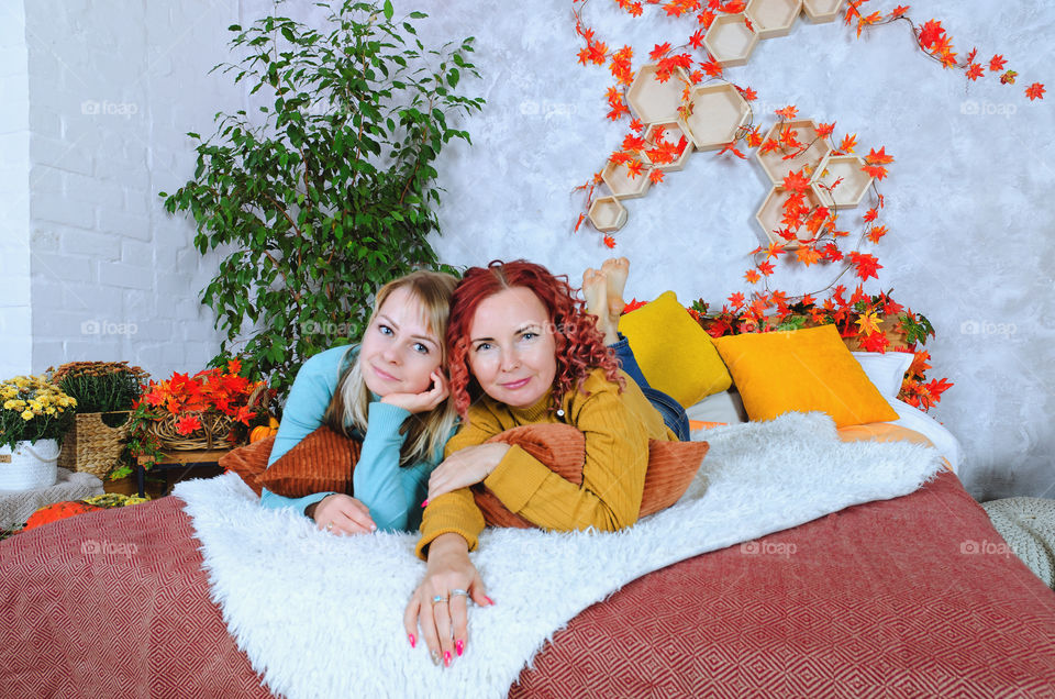 lovely women, sisters, friends in warm sweaters sitting in the bed at a cozy home, laughing, enjoying. Home interior with autumn decorations, orange leaves, flowers, pumpkins.