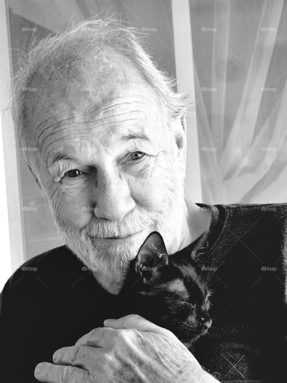 Black and white portrait of man holding a small black kitten.  Man is smiling. He is looking straight ahead. The kittens eyes are almost closed.  He is relaxed and happy to be held.