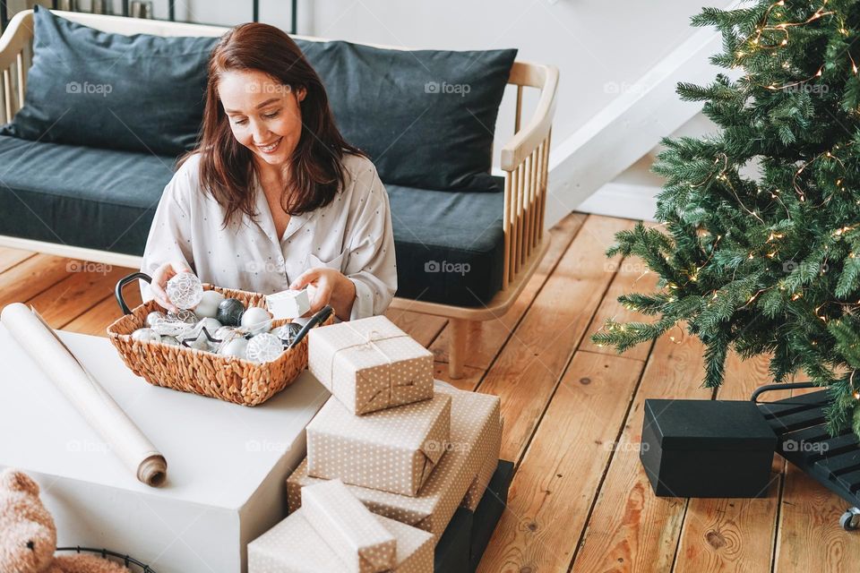 Young adult forty years woman with dark hair in cozy pajamas with present gift box sitting in living room with Christmas tree