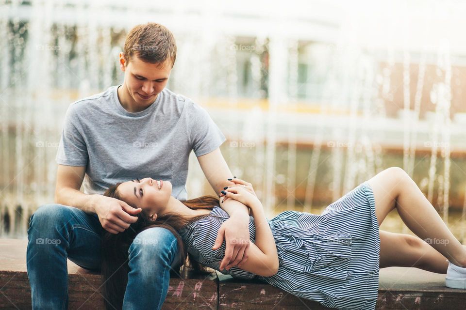 Happy young couple in love teenagers friends dressed in casual style sitting together near fountain on the city street