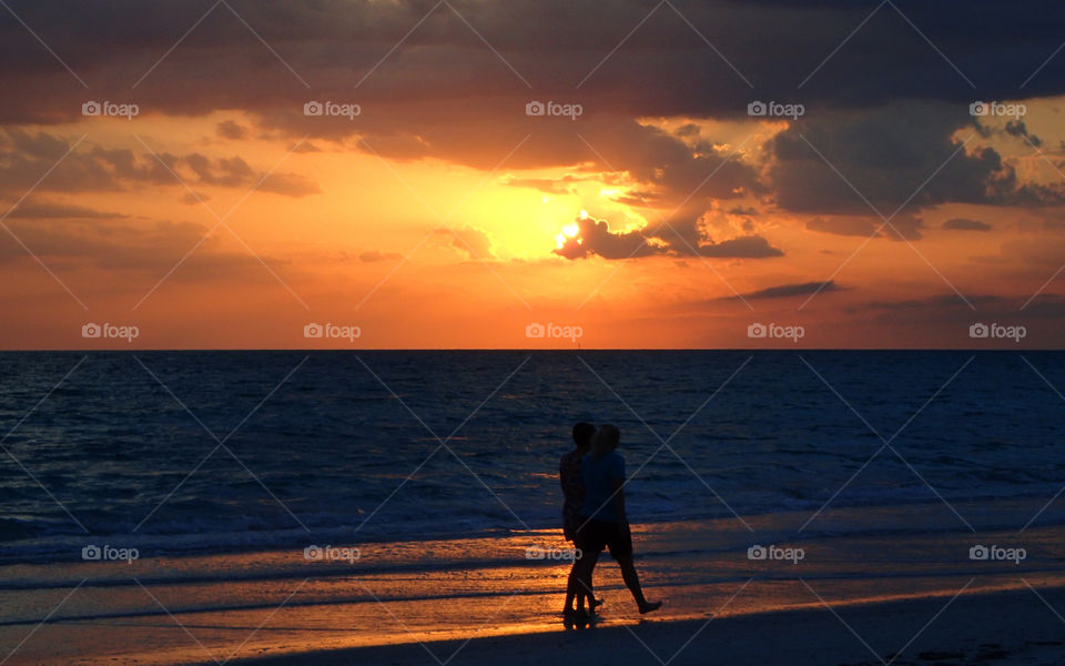 Couple strolling by seashore against a fiery golden summer sunset on the horizon