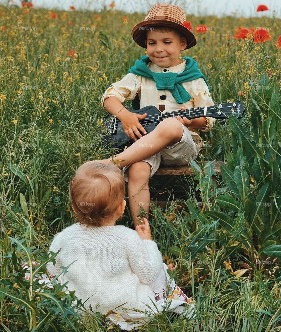 A toddler BOY playing on the guitar between flowers and a little girl listening