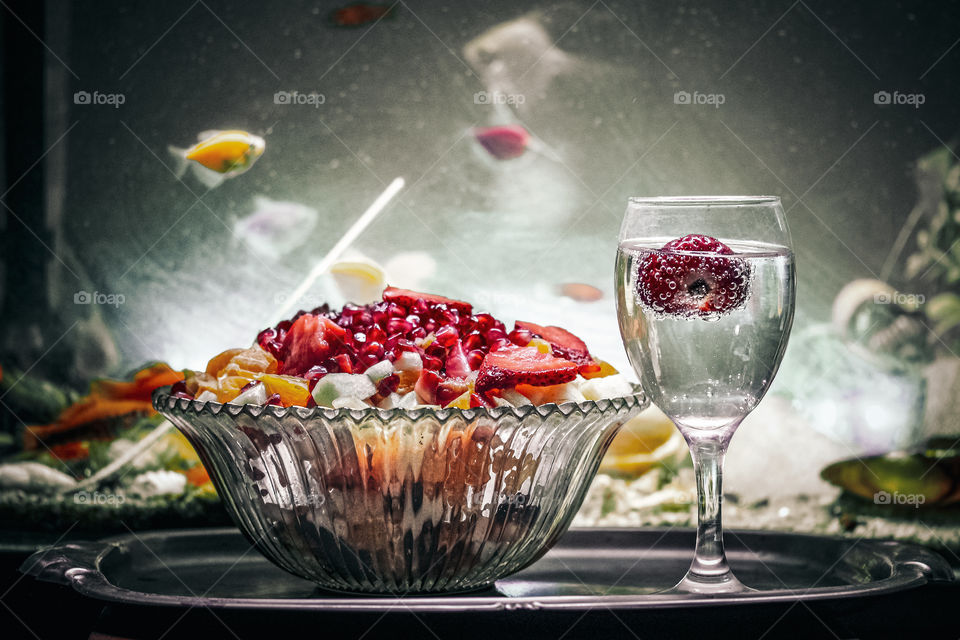 A fruit salad and the glass of mineral water with a strawberry at the glass. In the background there are aquarium fish swimming