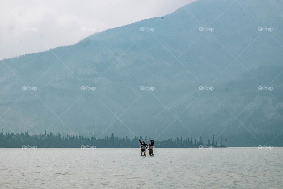 two people standing in front of mountain in shallow river near Canadian Rockies mountains in Alberta with blue water